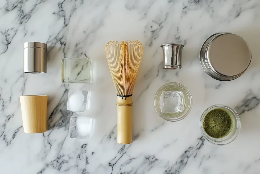 Flat lay of essential matcha tools, including a bamboo whisk, sifter, glass jar, and electric frother, arranged on a marble countertop for making iced matcha lattes.