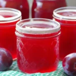 Homemade plum jelly in glass jars with a vibrant red color, surrounded by fresh plums on a green cloth. A visually appealing representation of a delicious plum jelly recipe.