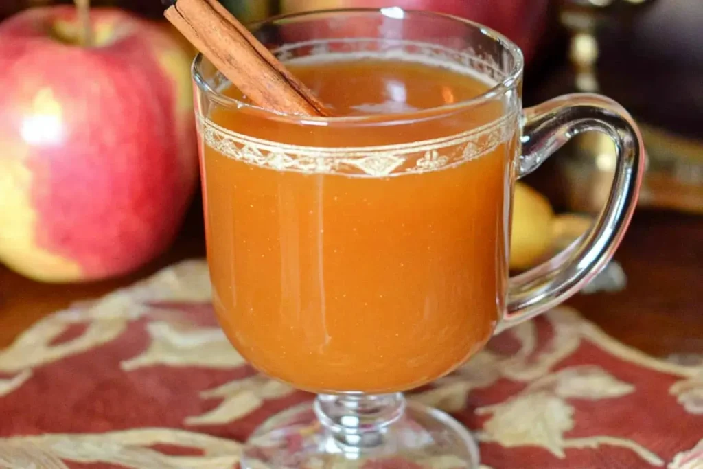 A clear glass mug of warm Fireball Apple Cider garnished with a cinnamon stick, sitting on a festive autumn-themed tablecloth with fresh apples in the background, capturing cozy fall vibes.