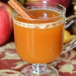 A clear glass mug of warm Fireball Apple Cider garnished with a cinnamon stick, sitting on a festive autumn-themed tablecloth with fresh apples in the background, capturing cozy fall vibes.