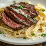 A gourmet dish of steak and pasta featuring medium-rare steak slices atop creamy fettuccine Alfredo, garnished with parsley and Parmesan, served on a white plate.